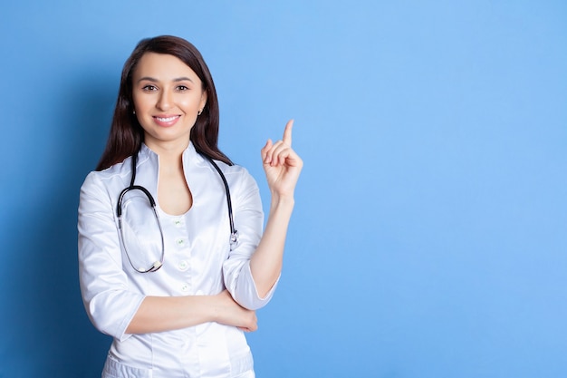 Portrait d'un beau jeune médecin d'une femme en robe blanche sur fond bleu Un endroit pour le texte et la publicité Pédiatre Thérapeute Médecin à domicile Médecin en ligne