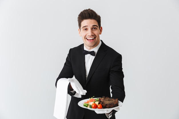 Portrait d'un beau jeune serveur en smoking montrant plat de steak de boeuf sur une assiette sur un mur blanc