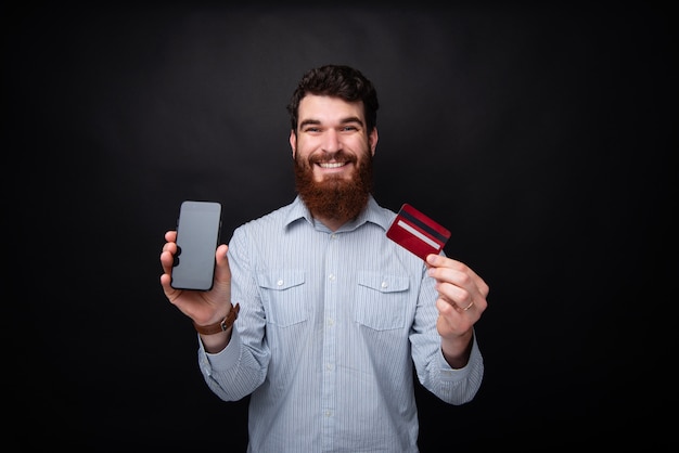 Portrait de beau mec barbu et à pleines dents souriant, montrant son téléphone portable et sa carte de crédit, debout sur un fond sombre isolé
