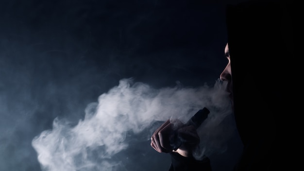 Portrait d'un beau mec dans un chapeau noir et des lunettes de soleil vaping et expirant un nuage de vapeur d'une cigarette électronique