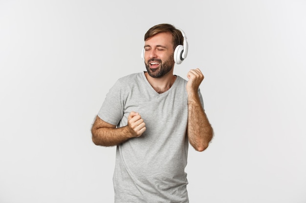 Portrait de beau mec heureux en t-shirt gris, écouter de la musique dans des écouteurs sans fil et danser