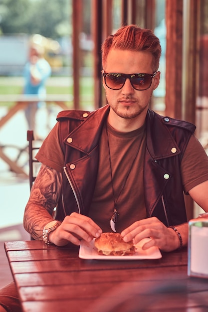 Portrait d'un beau mec hipster élégant assis à une table, commandé un hamburger et un soda, dîne dans un café en bordure de route