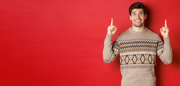 Portrait d'un beau mec rêveur pensant aux vacances de noël portant un pull d'hiver pointant et regardant l'offre promotionnelle du nouvel an debout sur fond rouge