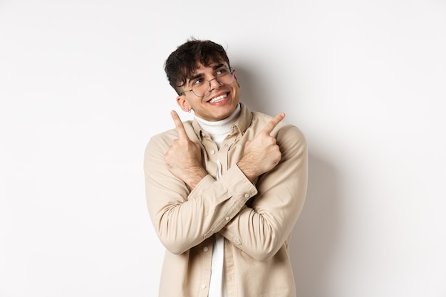 Portrait de beau mec souriant dans des verres pointant les doigts sur le côté, a fait son choix et regarde le logo du coin supérieur droit, debout sur le mur blanc.