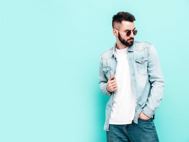 Portrait de beau modèle confiant hipster élégant hipster lambersexuel Homme vêtu d'une veste et d'un jean Mode homme posant près du mur bleu en studio en lunettes de soleil Réfléchi Isolé