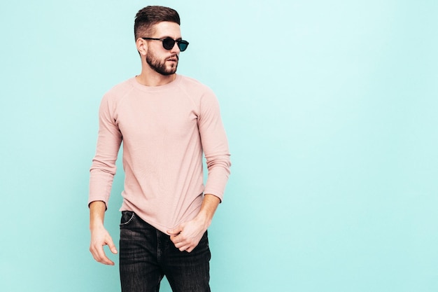 Portrait de beau modèle confiant Sexy homme élégant vêtu d'un pull rose et d'un jean Mode hipster mâle posant près du mur bleu en studio En lunettes de soleil Isolé