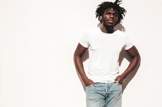 Portrait d'un beau modèle hipster Homme africain mal rasé vêtu d'un t-shirt d'été blanc et d'un jean Mode masculine avec une coiffure de dreadlocks posant près d'un mur blanc dans la rue