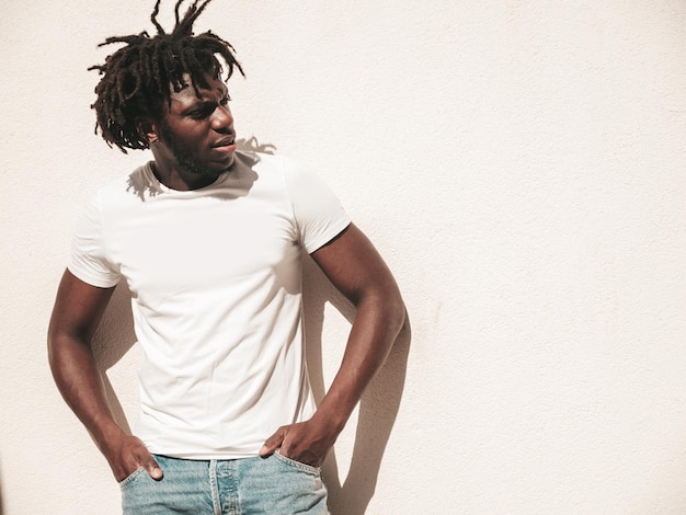 Portrait d'un beau modèle hipster souriant Un homme africain mal rasé vêtu d'un t-shirt d'été blanc et d'un jean Mode masculine avec une coiffure de dreadlocks posant sur le fond de la rue