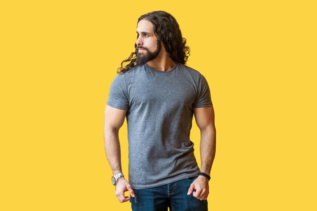 Portrait d'un beau modèle de jeune homme barbu brutal avec de longs cheveux bouclés en t-shirt gris debout et regardant loin avec un visage sérieux et fier tourné en studio à l'intérieur isolé sur fond jaune