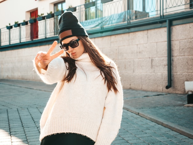 Photo portrait de beau modèle souriant. femme vêtue d'un pull et d'un bonnet blancs hipster chauds. elle pose dans la rue