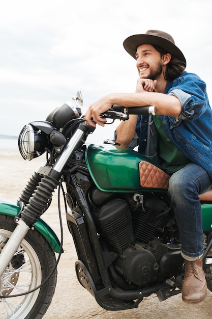 Portrait d'un beau motard optimiste joyeux sur son vélo à l'extérieur sur une plage portant un chapeau.