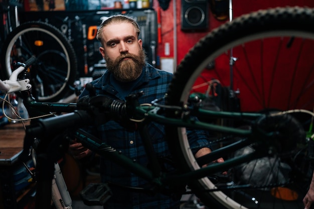 Portrait d'un beau réparateur cycliste barbu avec une coupe de cheveux de mode debout à vélo dans un atelier de réparation avec un intérieur sombre regardant la caméra