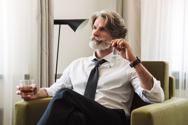 Portrait d'un beau rêve pensant homme d'affaires aux cheveux gris senior assis sur une chaise à l'intérieur à la maison buvant de l'alcool tenant des verres à la main.