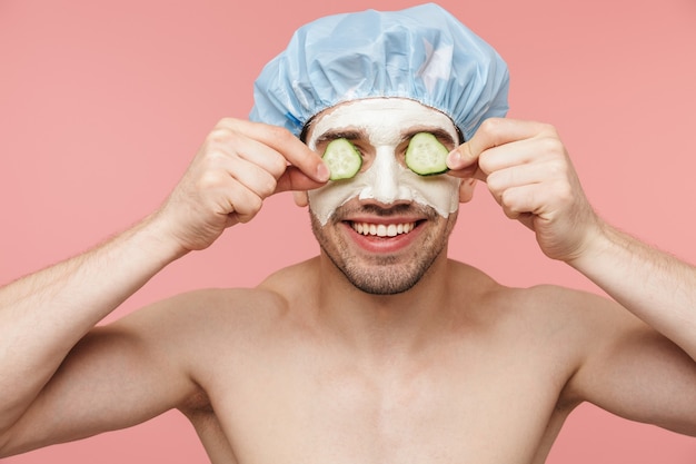 Portrait de beauté d'un bel homme brune torse nu souriant debout isolé sur un mur rose, portant un masque cosmétique de concombre facial