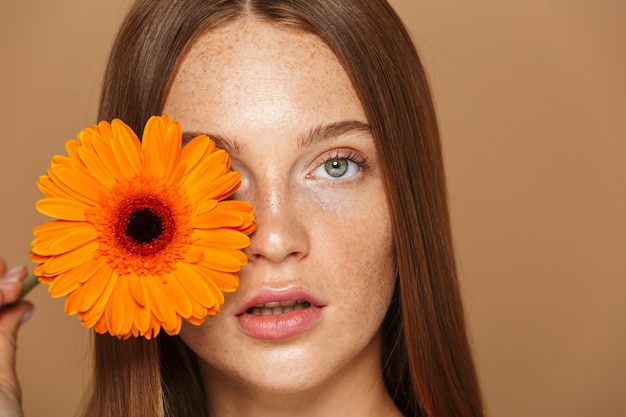 Portrait de beauté d'une belle jeune femme en bonne santé avec de longs cheveux rouges montrant fleur d'herbera orange isolé sur mur beige