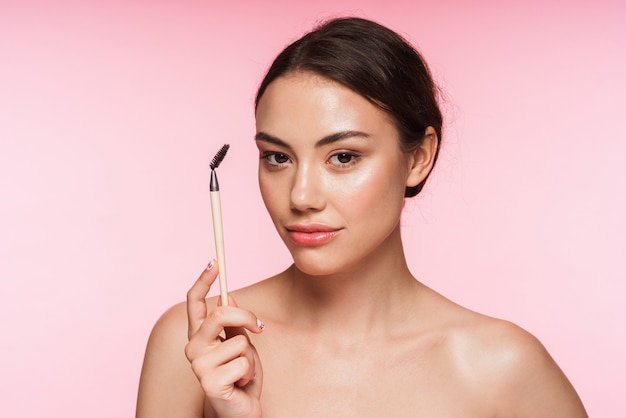 Portrait de beauté d'une belle jeune femme brune aux seins nus debout isolée sur rose, tenant une brosse à sourcils