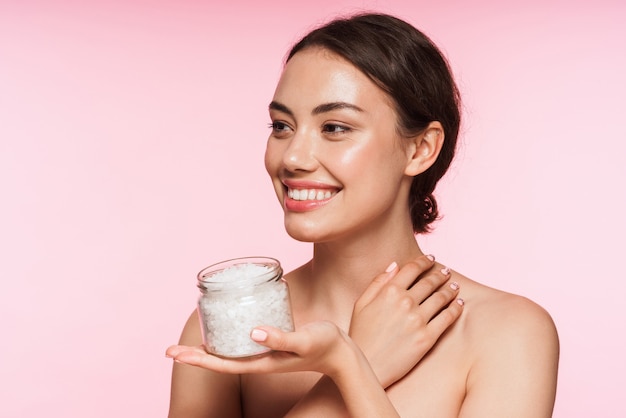 Portrait de beauté d'une belle jeune femme brune aux seins nus souriante debout isolée sur rose, montrant un pot avec une crème hydratante