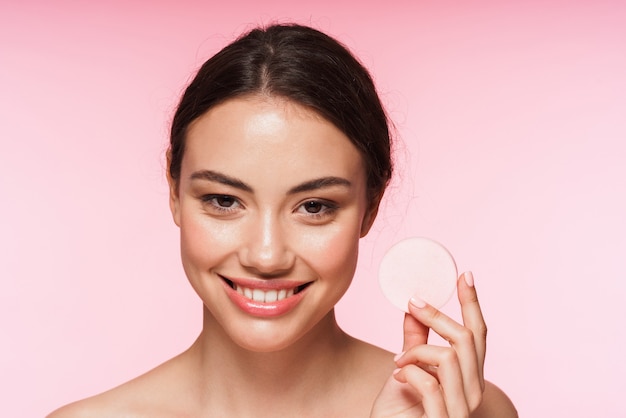 Portrait de beauté d'une belle jeune femme brune aux seins nus souriante debout isolée sur rose, tenant une éponge de maquillage sur son visage