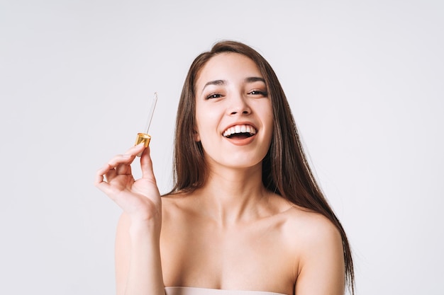 Portrait de beauté d'une femme asiatique souriante et heureuse aux cheveux longs noirs avec une pipette à l'huile biologique sur le visage et les mains de la peau propre et fraîche sur fond blanc isolé