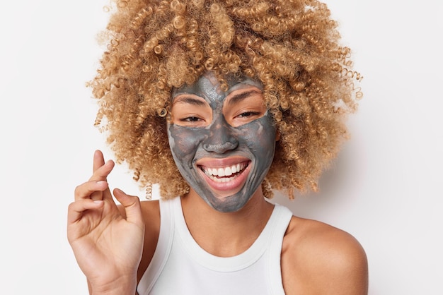 Le portrait de beauté d'une femme aux cheveux bouclés heureuse applique un masque hydratant sur le visage sourit largement montre des dents blanches porte des poses de t-shirt décontractées à l'intérieur. Concept de soins de la peau et de rajeunissement de bien-être.