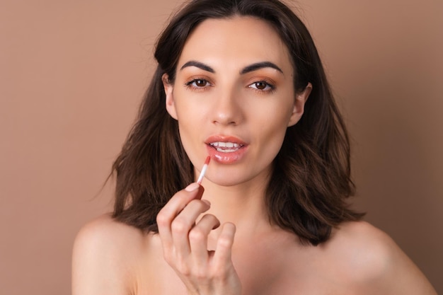 Photo portrait de beauté d'une femme aux seins nus avec une peau parfaite et un maquillage naturel sur fond beige tenant un rouge à lèvres brillant au chocolat brun