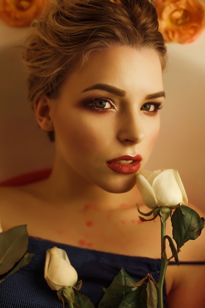 Portrait de beauté d'une femme blonde séduisante avec un maquillage parfait prend un bain avec de l'eau rouge et des roses blanches