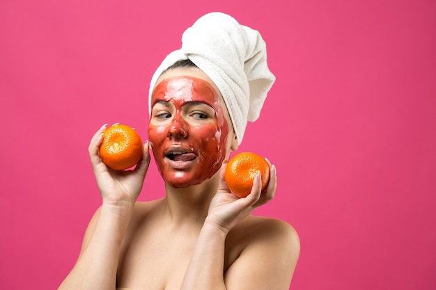 Portrait de beauté d'une femme dans une serviette blanche sur la tête avec un masque nourrissant rouge sur le visage Soins de la peau nettoyage spa cosmétique bio bio concept de détente Une fille se tient le dos tenant une mandarine orange