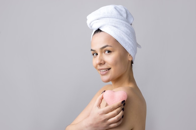 Portrait de beauté de femme en serviette blanche sur la tête avec une éponge pour un corps en vue d'un coeur rose. Concept de détente de spa cosmétique bio écologique de nettoyage de soins de la peau.