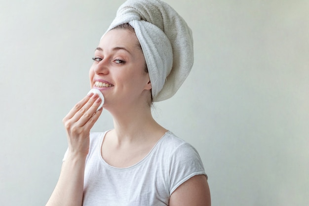 Portrait de beauté de femme souriante dans une serviette sur la tête avec une peau douce et saine enlever le maquillage avec un coton isolé sur fond blanc.