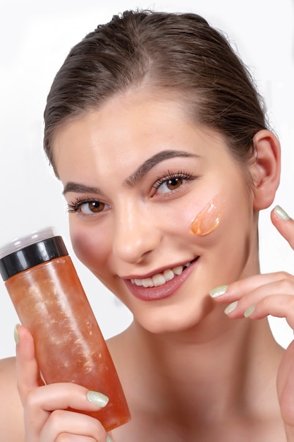 Portrait de beauté en gros plan d'une belle femme riante appliquant une crème pour le visage et regardant isolée