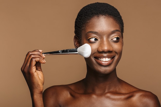 Portrait de beauté d'une jeune femme africaine à moitié nue heureuse tenant un pinceau de maquillage isolé sur beige
