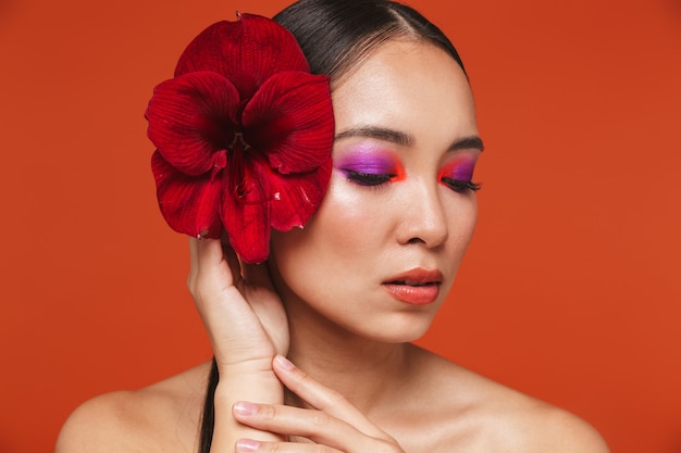 Portrait de beauté d'une jeune femme asiatique aux seins nus aux cheveux bruns portant un maquillage lumineux, debout isolée sur rouge, posant avec une fleur rouge