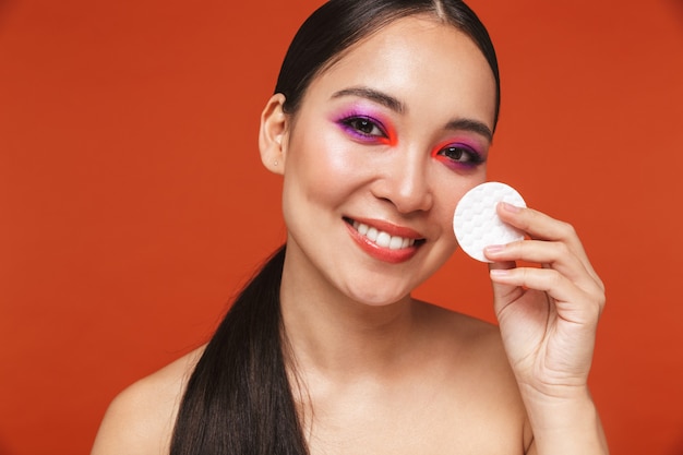 Portrait de beauté d'une jeune femme asiatique aux seins nus heureuse avec des cheveux bruns portant un maquillage lumineux, debout isolée sur rouge, à l'aide de cotons