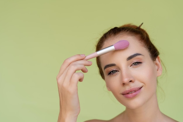 Portrait de beauté d'une jeune femme aux cheveux roux aux seins nus avec des épaules nues sur fond vert avec une peau parfaite et un maquillage naturel tient un pinceau blush