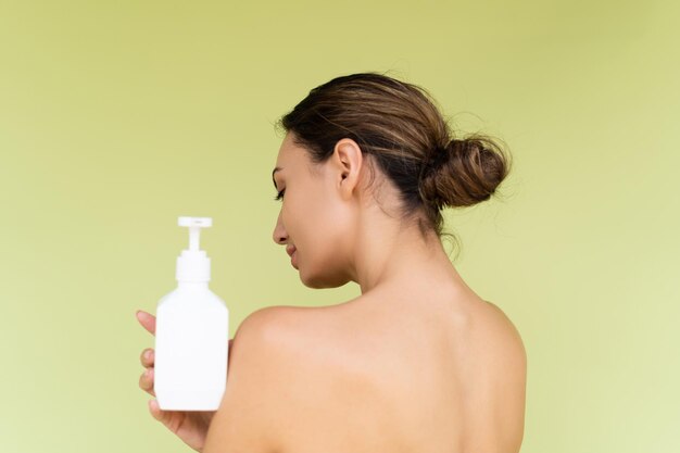 Portrait de beauté d'une jeune femme aux seins nus avec des épaules nues sur fond vert avec une peau parfaite et un maquillage naturel tenant une bouteille de lotion pour le corps de shampooing