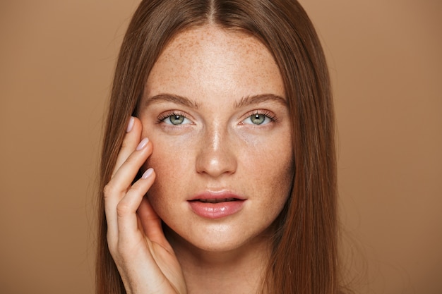 Portrait de beauté d'une jeune femme aux seins nus sensuelle avec de longs cheveux rouges posant, tenant la main sur son visage isolé sur mur beige