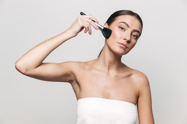 Portrait de beauté d'une jeune femme brune attrayante en bonne santé debout isolé, appliquant le maquillage avec un pinceau