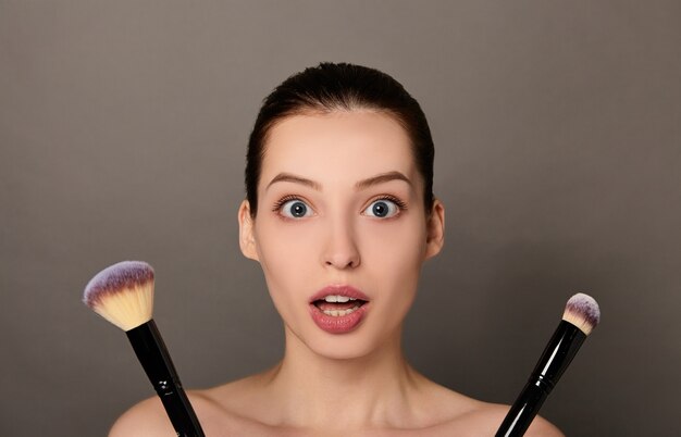 Portrait de beauté d'une jeune femme surprise tenant deux pinceaux de maquillage sur fond marron clair