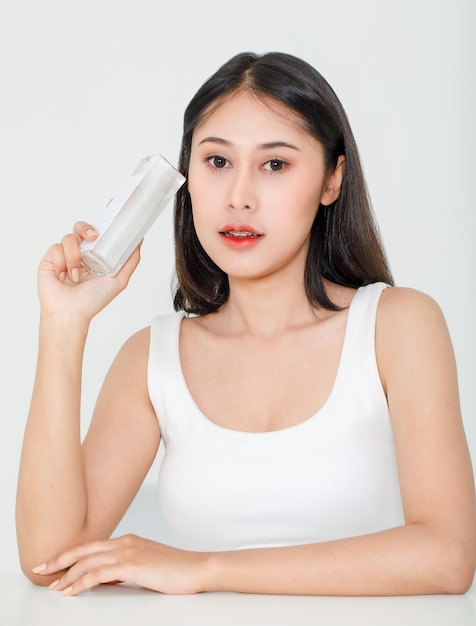 Portrait de beauté d'un jeune mannequin asiatique aux cheveux noirs courts en maillot de corps débardeur tenant un paquet de bouteille de soin de la peau à la crème pour le visage au sérum naturel à la main sur fond blanc.