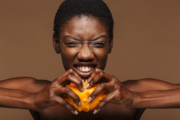 Portrait de beauté d'une jolie jeune femme africaine à moitié nue tenant un melon à cornes kiwano isolé sur marron