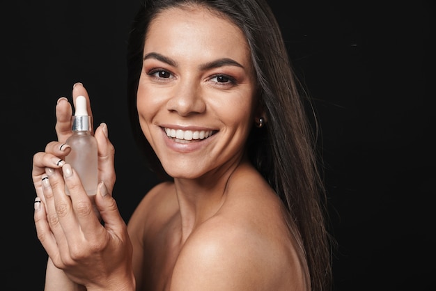 Portrait de beauté d'une jolie jeune femme aux seins nus avec de longs cheveux bruns isolés sur un mur noir, montrant de l'huile de beauté de maquillage