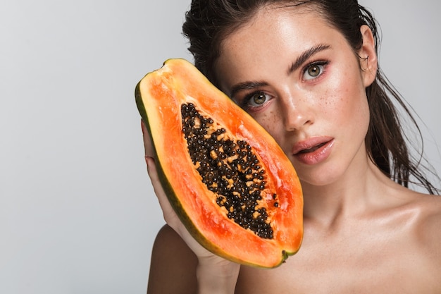 Portrait de beauté d'une jolie jeune femme brune seins nus debout isolé sur blanc, posant avec papaye