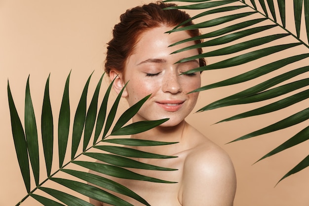 Portrait de beauté d'une jolie jeune femme rousse topless debout isolée, posant entourée de feuilles tropicales