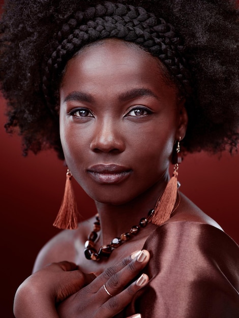 Portrait de beauté naturelle et femme noire à la mode en studio isolé sur fond rouge Cosmétiques de maquillage pour le visage et modèle africain sérieux avec tenue élégante bijoux traditionnels ou esthétique