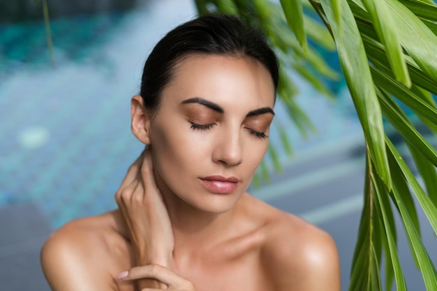 Portrait de beauté peau douce Modèle féminin avec maquillage naturel et peau saine derrière une plante à feuilles vertes Portrait d'une belle fille avec des ongles nus épaules nues