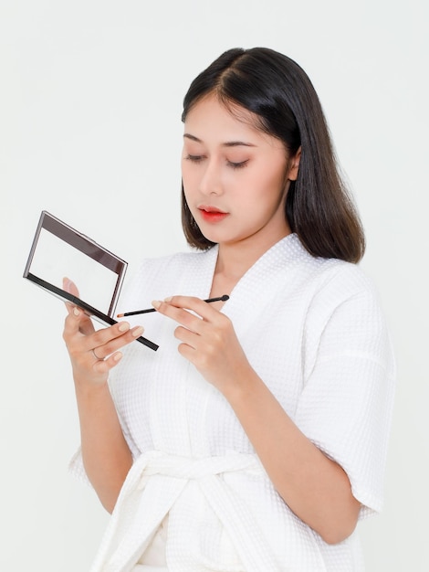 Portrait de beauté photo d'une jeune mannequin asiatique millénaire portant du maquillage dans une corde de douche debout souriant tenant une ombre à paupières colorée avec miroir et pinceau sur fond blanc.