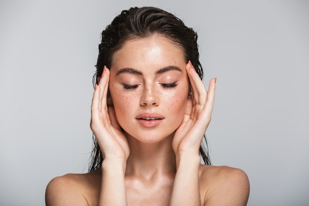 Portrait de beauté d'une séduisante jeune femme sensuelle souriante aux cheveux longs brune mouillée, isolée sur fond gris, touchant son visage, posant