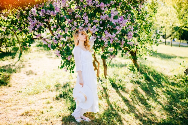 Portrait de beaux-arts de jeune fille blonde en robe élégante blanche en été lilas jardin fleuri. Buissons colorés avec des fleurs sur les branches.