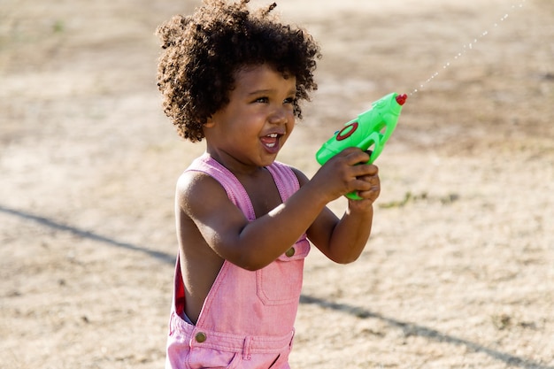 Portrait d'un bébé afro-américain jouant avec un pistolet à eau dans le parc.