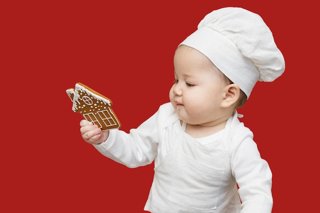 Un portrait d'un bébé asiatique dans une toque et un tablier regardant un biscuit au gingembre de Noël Isoler sur un fond vineux